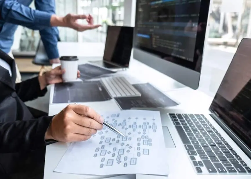 Two developers work at a bright desk with code on a large monitor, a laptop, printed diagrams, and a coffee cup nearby.