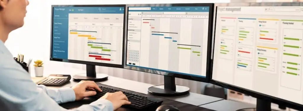 Person at a desk using three monitors that display project management dashboards and Gantt charts, tracking timelines and tasks.