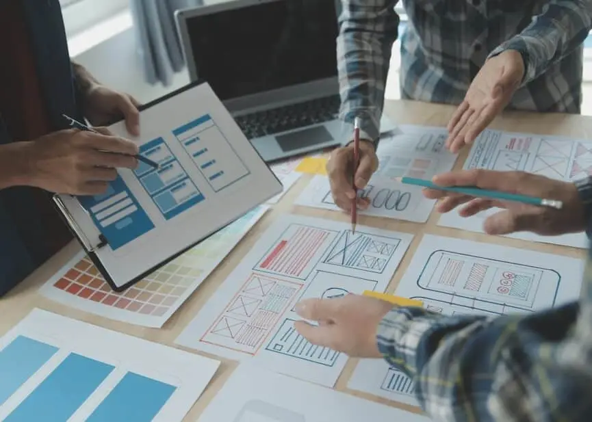 Team collaborates around a table, sketching UI wireframes on paper with pens, while a tablet and laptop lay nearby for reference.