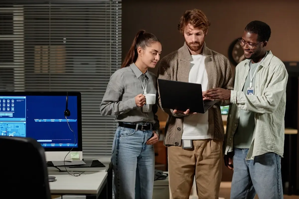 Three coworkers stand around a laptop in an office, discussing a project; one holds a mug.