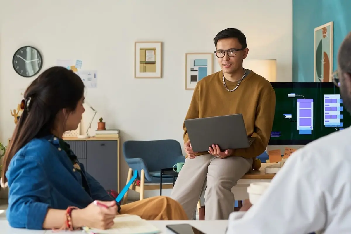 Male presenter in a brown sweater shares a laptop briefing with two colleagues in a modern office.