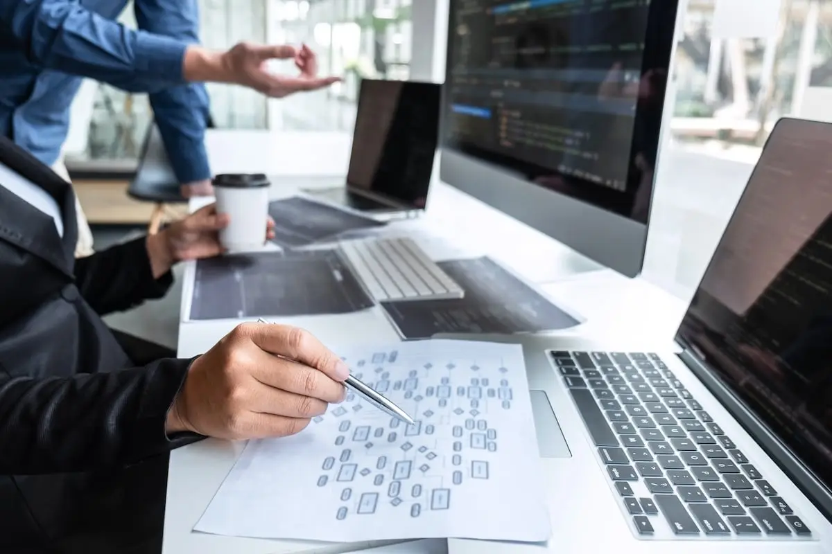 Two developers work at a bright desk with code on a large monitor, a laptop, printed diagrams, and a coffee cup nearby.