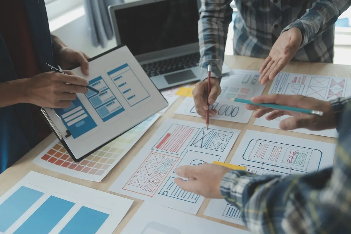 Team collaborates around a table, sketching UI wireframes on paper with pens, while a tablet and laptop lay nearby for reference.