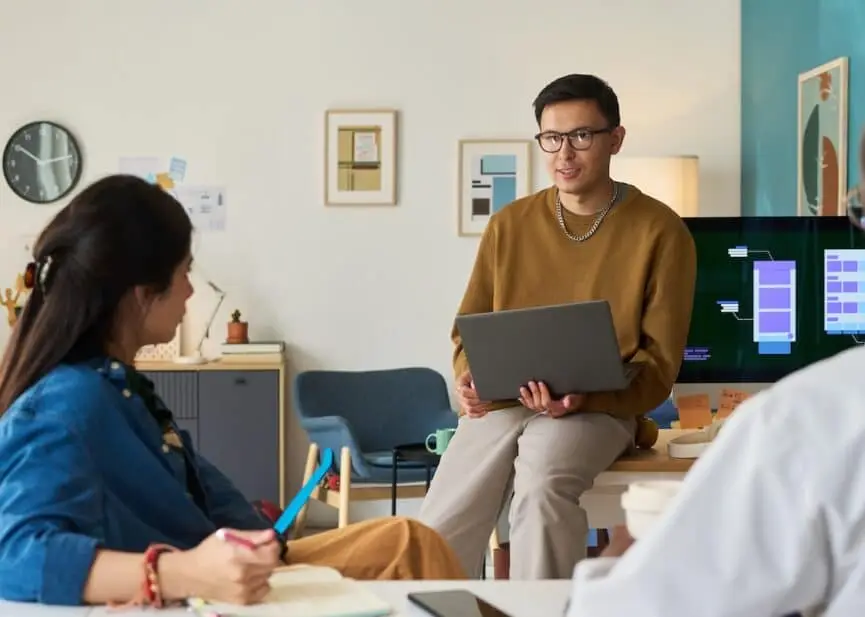 Male presenter in a brown sweater shares a laptop briefing with two colleagues in a modern office.